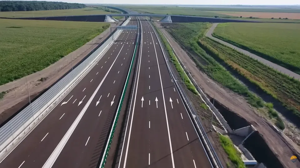 O autostradă cu mai multe benzi de rulare, marcaje albe și bariere metalice de siguranță traversează un peisaj de câmpie verde sub un cer deschis.