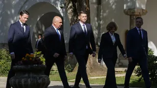 Cinci persoane îmbrăcate în costume închise de culoare merg pe un drum pietruit lângă vegetație și clădiri.