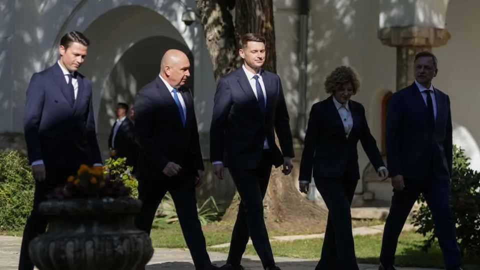 Cinci persoane îmbrăcate în costume închise de culoare merg pe un drum pietruit lângă vegetație și clădiri.