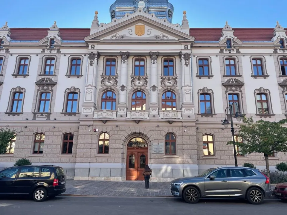 O clădire monumentală cu fațadă decorată, ferestre multiple și un fronton central, cu două mașini parcate în prim-plan pe un drum asfaltat.