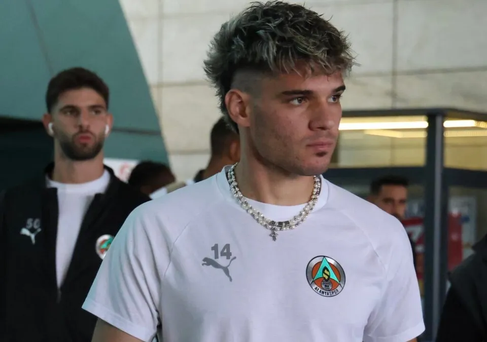 Un bărbat cu părul scurt și șaten poartă un tricou alb cu un logo circular și numărul 14, stând lângă un alt bărbat într-o vestă neagră.