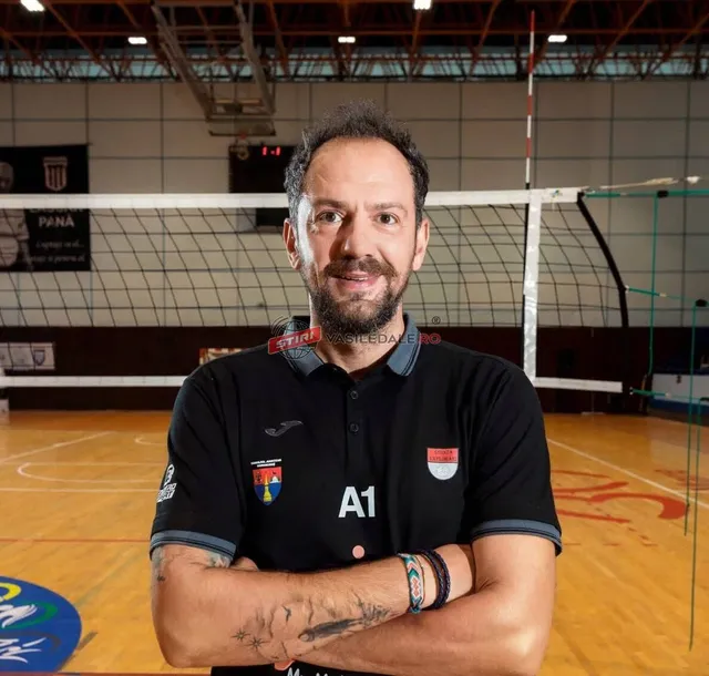 Un bărbat cu barbă și păr scurt stă cu brațele încrucișate într-o sală de sport cu un teren de volei în fundal.