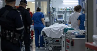 Trei persoane în uniforme medicale și doi polițiști stau în jurul unui pat de spital acoperit cu un cearșaf alb într-un mediu clinic.
