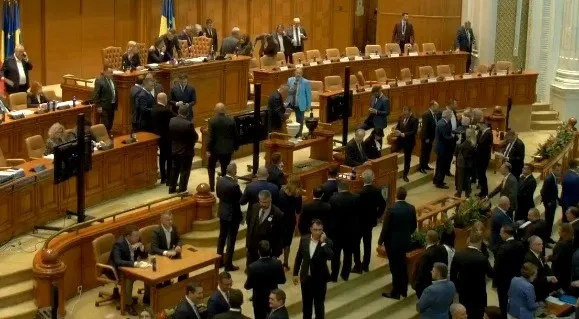 Oamenii în costume purtând haine de culoare închisă stau sau merg într-o sală mare cu mese de lemn și scaune dispuse în rânduri.