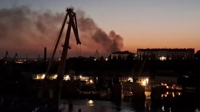 Un turn de ridicare se ridică în siluetă pe fundalul unui cer la apus, deasupra unui port cu clădiri și fum în depărtare.