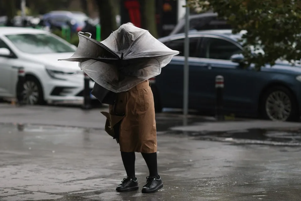 O persoană cu o fustă maro și pantofi negri merge pe un trotuar umed ținând un umbrelă gri întoarsă în vânt.