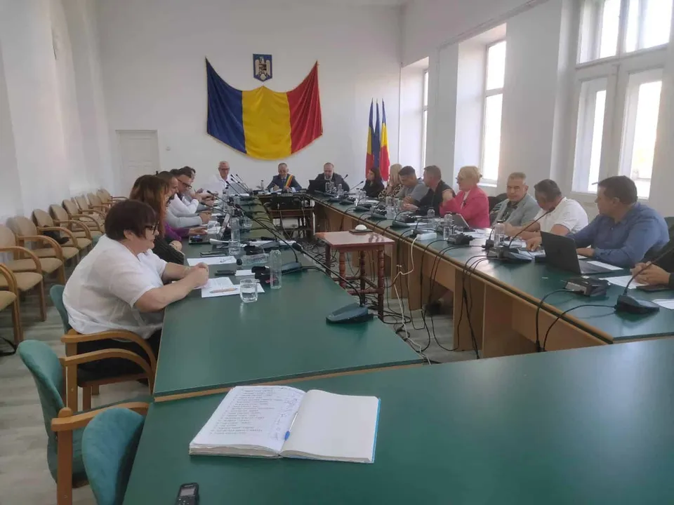 Oamenii stau la mese lungi cu suprafețe verzi într-o sală de ședințe, sub un drapel tricolor afișat pe perete.