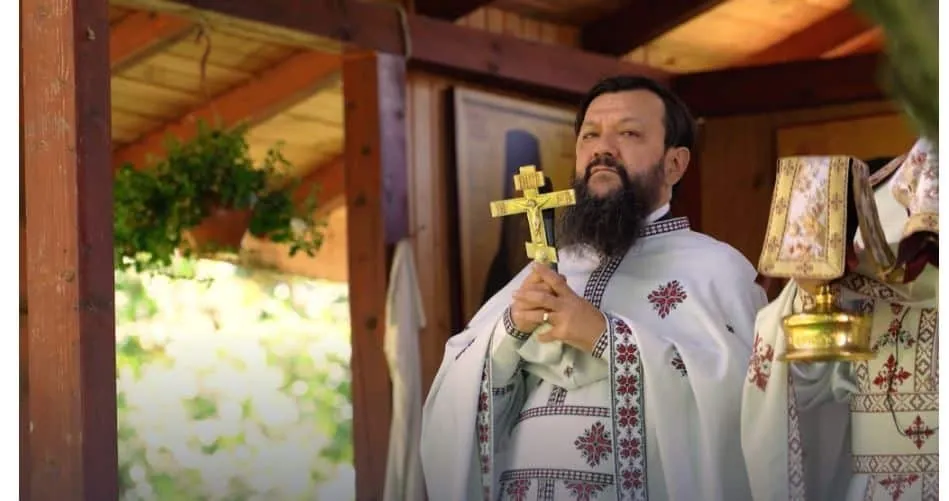 Un bărbat cu barbă poartă o robă albă decorată și ține un crucifix de aur în mâna dreaptă.