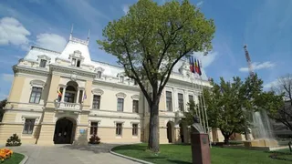 O clădire de stil clasic cu ferestre și balcoane este situată lângă un copac mare și un bust pe un soclu sub un cer albastru.