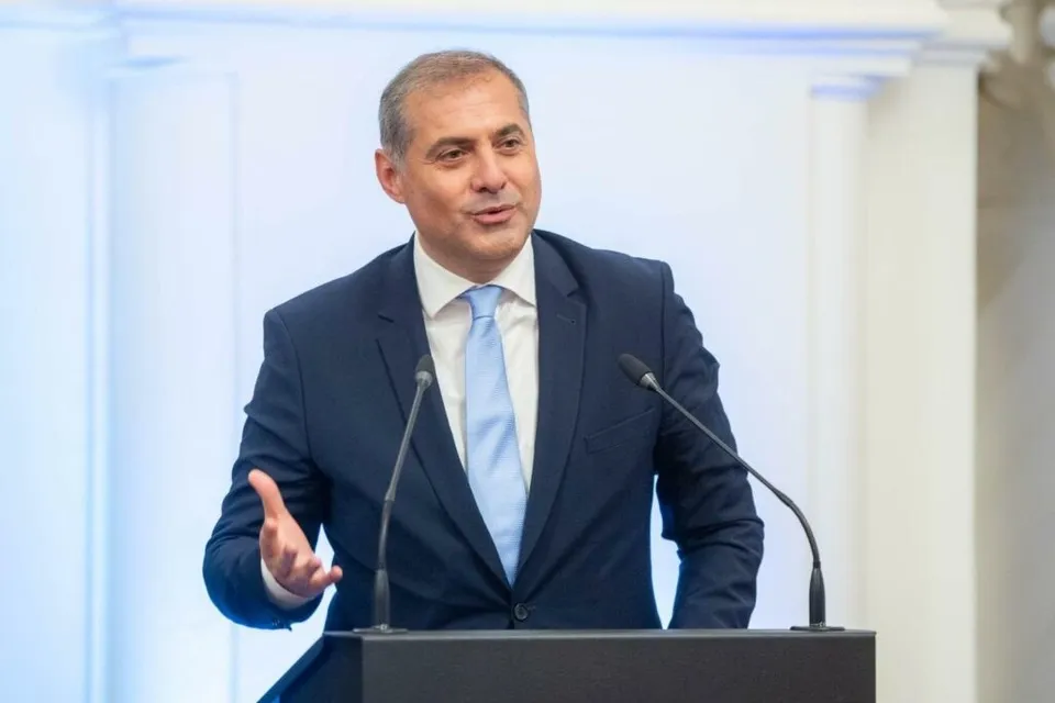 Un bărbat într-un costum închis la culoare și cravată albastră stă în spatele unui podium cu două microfoane.