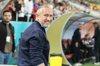 Un bărbat cu părul gri și bluza albastră zâmbește, fiind poziționat în fața unui banc de substituție într-un stadion.
