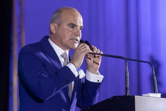 Un bărbat cu părul scurt și costum albastru vorbește la un microfon aflat în fața unui podium.