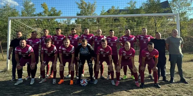 Un grup de bărbați în echipamente sportive de culoare roșie și neagră pozează în fața unei porți de fotbal pe un teren de iarbă.