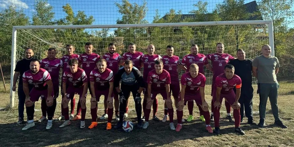 Un grup de bărbați în echipamente sportive de culoare roșie și neagră pozează în fața unei porți de fotbal pe un teren de iarbă.