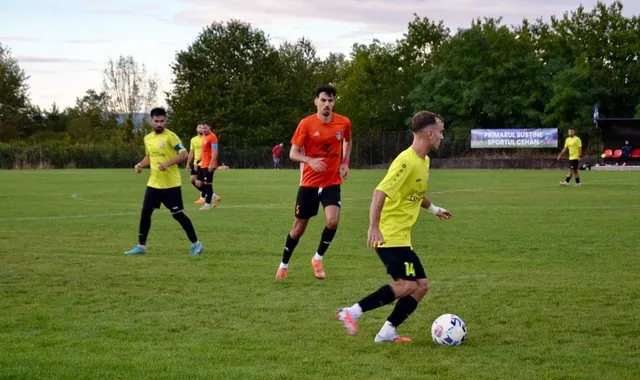 Câțiva jucători de fotbal de pe un teren de iarbă verde sunt în mișcare, unul având un balon la picioare.
