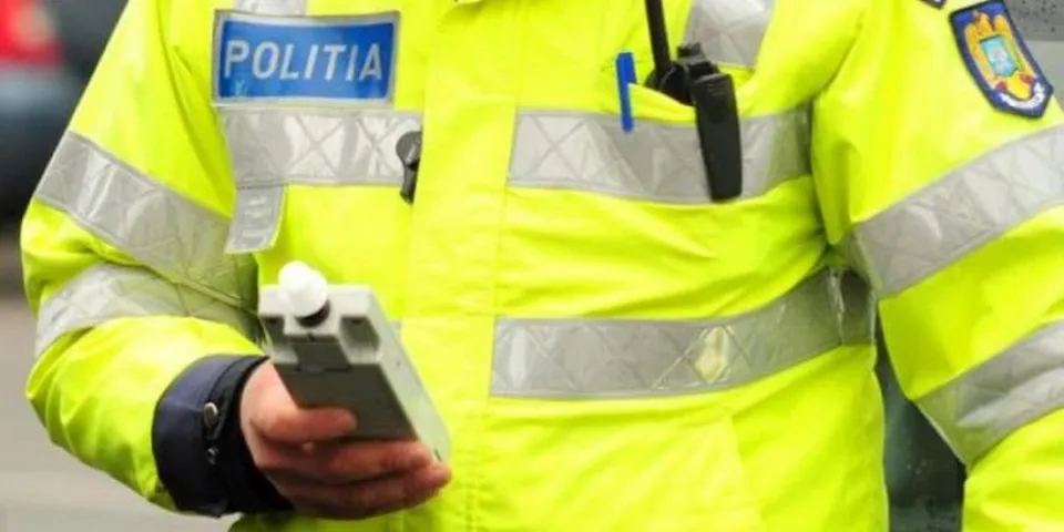 Un polițist în uniformă galbenă fluorescentă cu inscripția 'POLITIA' ține un etilotest electronic gri în mână.