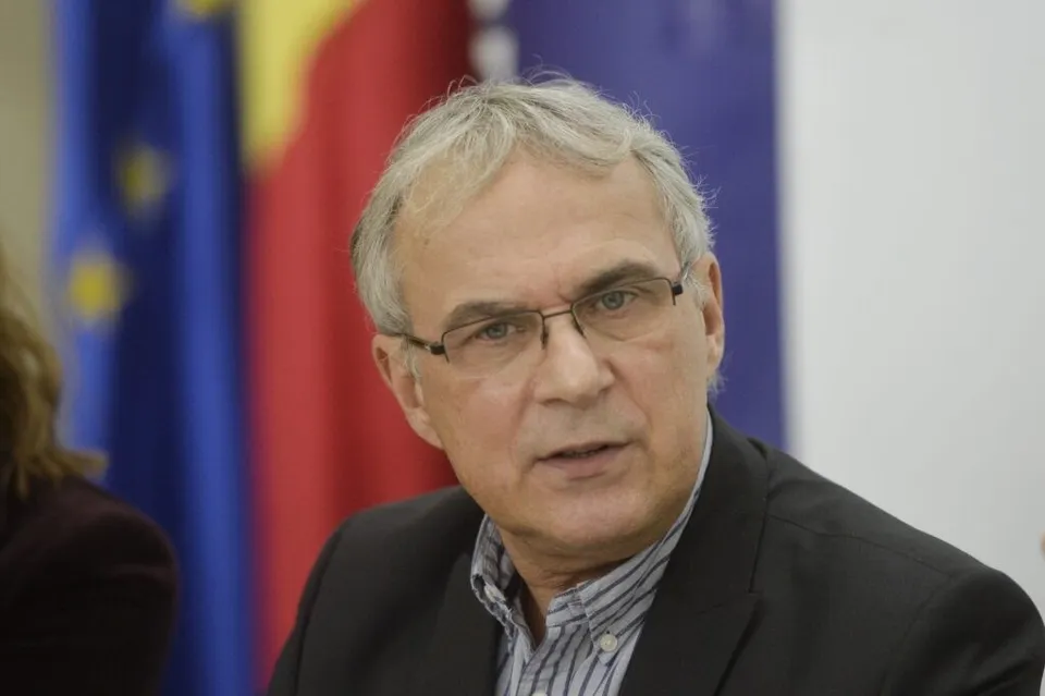 Un bărbat cu ochelari și părul cărunt poartă o jachetă neagră peste o cămașă în dungi, fiind poziționat în fața unor steaguri colorate.