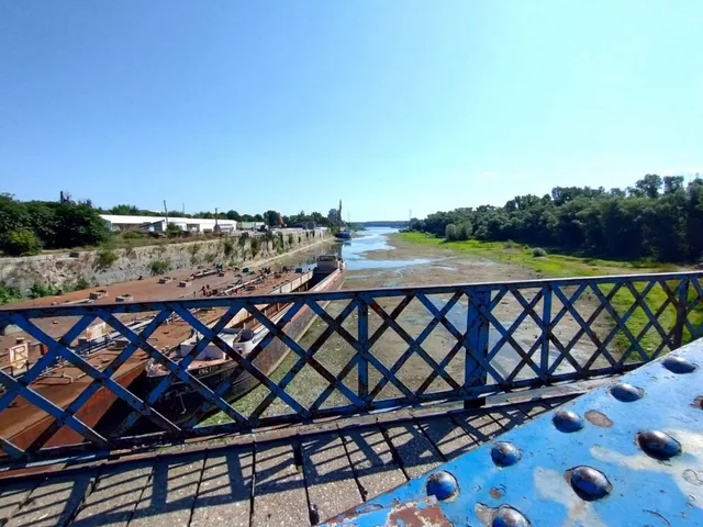 O privire de sus asupra unui râu cu apă puțină, alături de nave mari și un pod cu balustradă metalică albastră.