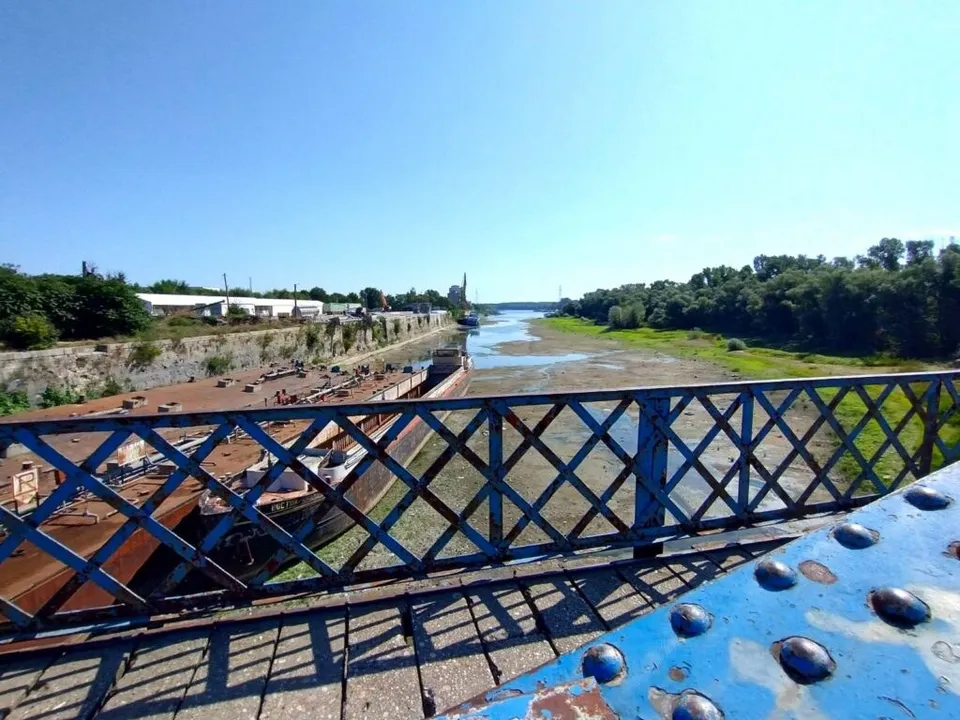 O privire de sus asupra unui râu cu apă puțină, alături de nave mari și un pod cu balustradă metalică albastră.