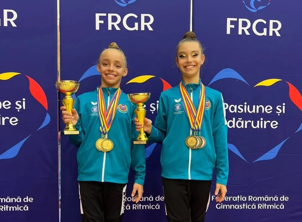 Două fete zâmbitoare poartă jachete verzi și medalii aurii, ținând trofee aurii în mâini în fața unui fundal albastru cu inscripții albe.