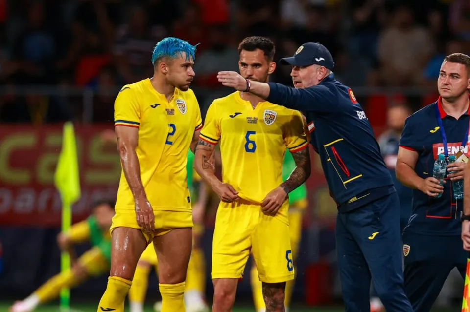 Trei bărbați stau pe un teren de fotbal, doi purtând echipamente galbene și unul purtând o șapcă și un costum sportiv albastru.