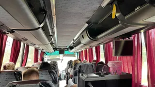 Interiorul unui autobuz cu pasageri așezați pe scaune, având draperii roșii la ferestre și un ecran suspendat pe tavan.
