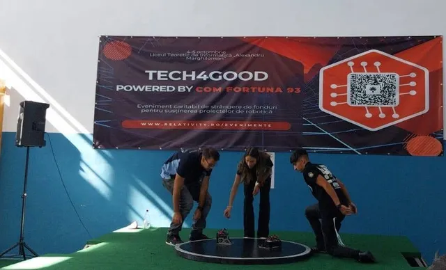 Trei persoane stau aplecate peste o platformă circulară pe o scenă verde în fața unui panou informativ mare cu textul Tech4Good.