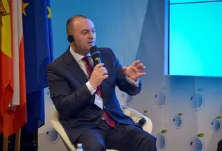 Un bărbat într-un costum închis și cravată roșie vorbește într-un microfon, stând așezat pe un scaun în fața unor steaguri.