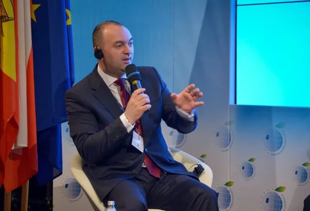 Un bărbat într-un costum închis și cravată roșie vorbește într-un microfon, stând așezat pe un scaun în fața unor steaguri.