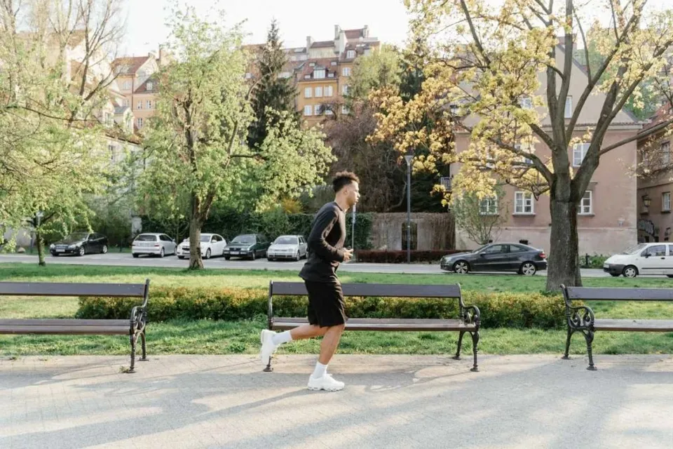Un bărbat într-un tricou negru și pantaloni scurți negri aleargă pe un drum pavat lângă niște bănci de parcă și copaci.