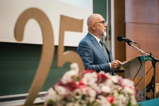 Un bărbat cu ochelari și barbă căruntie, purtând un costum albastru, vorbește la un podium lângă un număr mare de culoarea aurului.