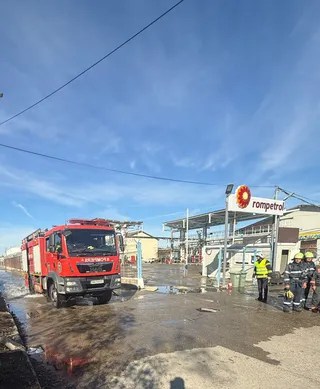Un camion de pompieri roșu stă pe un teren umed lângă un grup de persoane purtând echipamente de protecție galben și negru sub un cer senin.