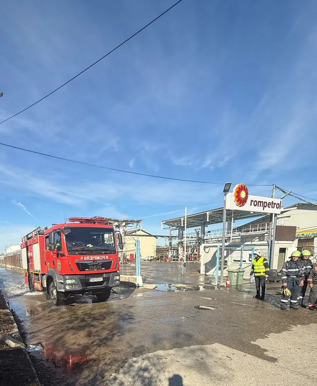 Un camion de pompieri roșu stă pe un teren umed lângă un grup de persoane purtând echipamente de protecție galben și negru sub un cer senin.