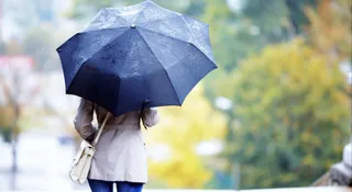 O persoană văzută din spate ține o umbrelă neagră, purtând palton deschis, pantaloni albaștri și poșetă deschisă.