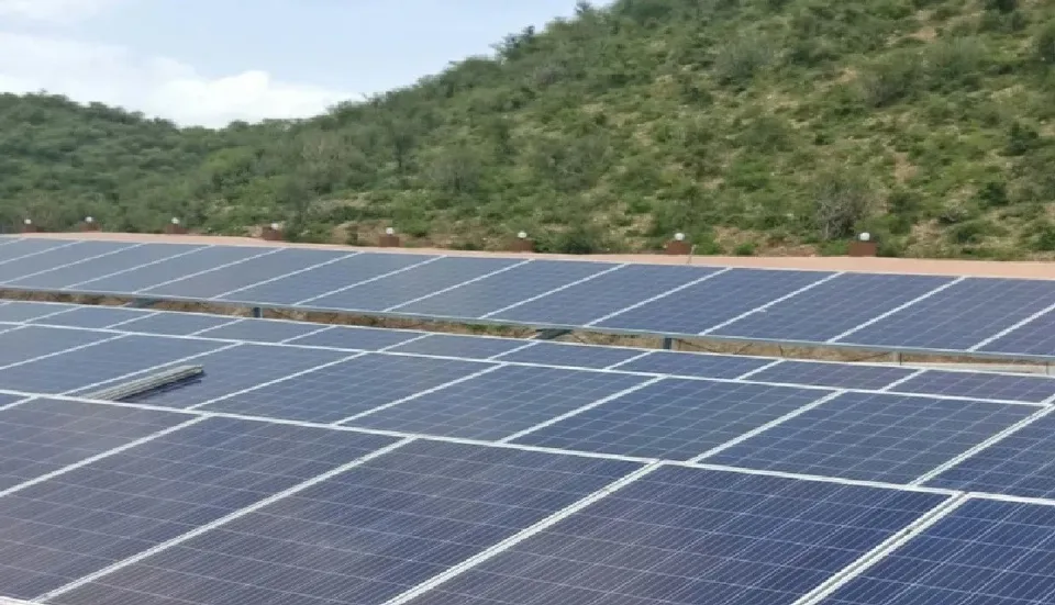 Rânduri de panouri solare sunt instalate pe un teren plat, având un deal împădurit în fundal sub un cer înnorat.
