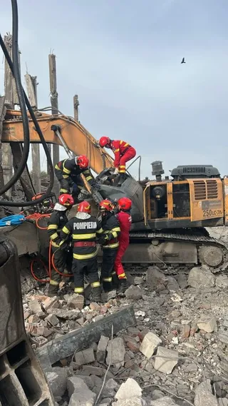 Mai mulți oameni purtând echipamente de protecție și casci roșii stau lângă un excavator galben aflat pe un teren cu resturi de beton și pietre.