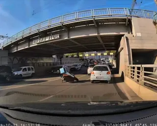 O motocicletă este înclinată pe asfaltul unei străzi sub un pasaj de beton, în apropierea unor mașini parcate sau în trafic.