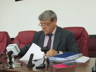 Un bărbat cu ochelari și sacou gri stă la o masă de lucru, citind un document alb lângă microfoane și un telefon mobil.