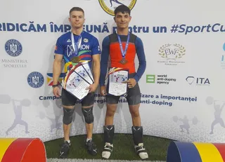 Doi bărbați în costume de halterofil stau în picioare purtând medalii și certificate în fața unui panou alb cu diverse logouri.