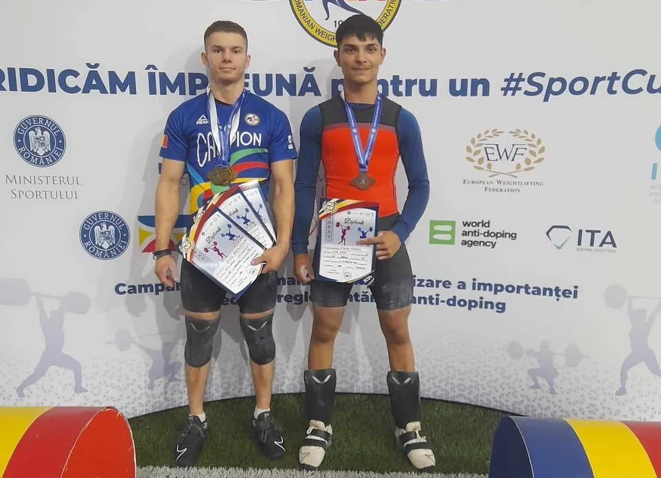 Doi bărbați în costume de halterofil stau în picioare purtând medalii și certificate în fața unui panou alb cu diverse logouri.