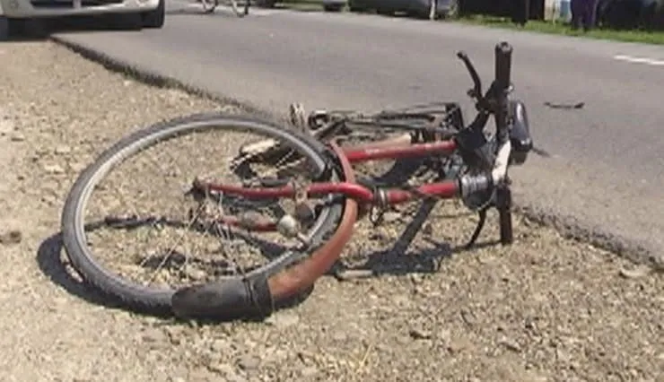 O bicicletă roșie cu roți de culoare închisă este întinsă pe marginea unui drum asfaltat lângă un teren cu pietriș.