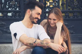 Un bărbat cu tatuaje pe antebraț și o femeie cu părul lung stau împreună și zâmbesc în fața unui gard metalic decorat.