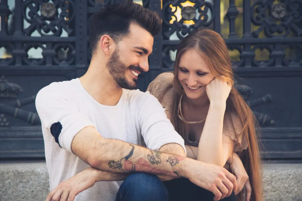 Un bărbat cu tatuaje pe antebraț și o femeie cu părul lung stau împreună și zâmbesc în fața unui gard metalic decorat.
