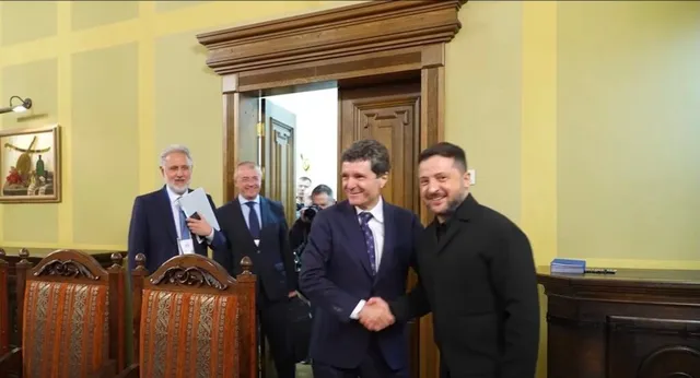 Patru bărbați în costume și haine închise stau în interiorul unei încăperi cu pereți galbeni și uși din lemn.