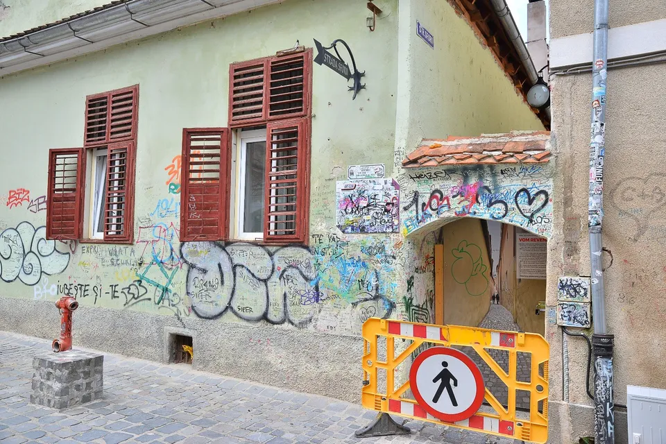 O clădire cu ferestre maronii și graffiti pe perete este vizibilă lângă un obstacol stradal portocaliu cu un semn de pieton.