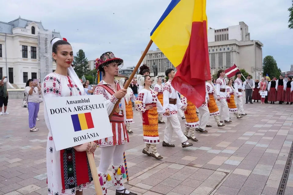 O grupă de persoane purtând costume populare tradiționaleionale merge într-un rând pe o piață pavată, un bărbat ridicând un steag galben, albastru și roșu.