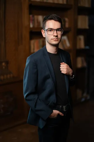 Un bărbat cu ochelari, purtând sacou albastru și tricou negru, stă în fața unor rafturi cu cărți într-un decor cu mobilier din lemn.
