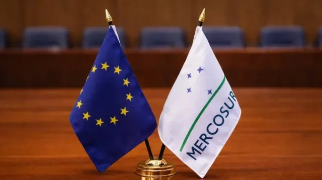 Două steaguri mici, unul albastru cu stele galbene și unul alb cu textul MERCOSUR și linii verzi, sunt așezate pe o suprafață de lemn.