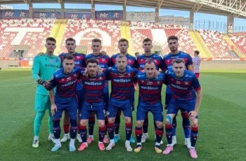 O echipă de fotbaliști în echipament sportiv albastru și roșu stă poziționată pe un teren de fotbal verde în fața unor tribune colorate.