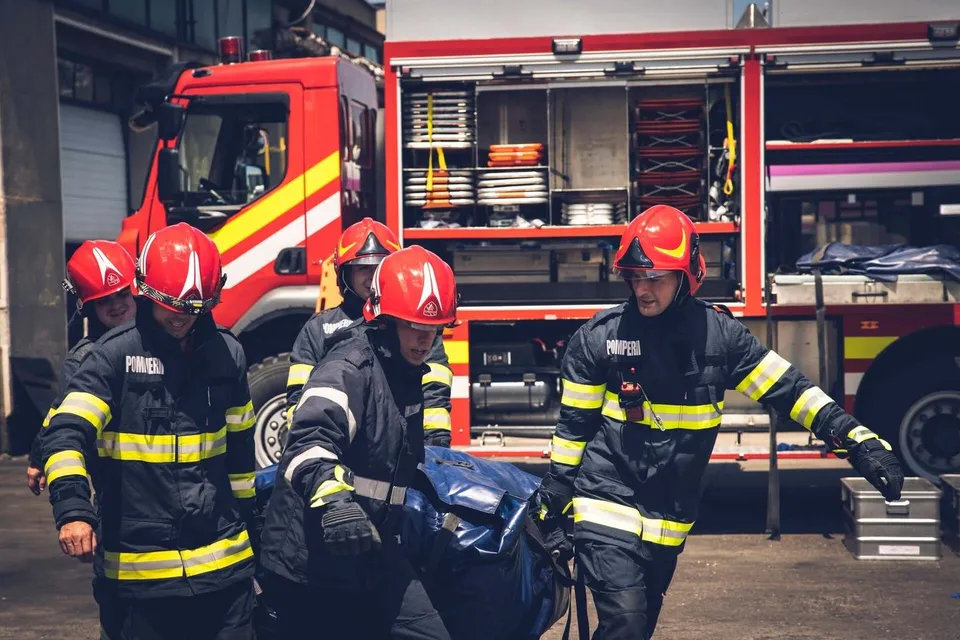 Patru pompieri în uniforme de protecție cu benzi reflectorizante transportă un echipament sac de culoare albastră lângă un autospecială de stingere a incendiilor.